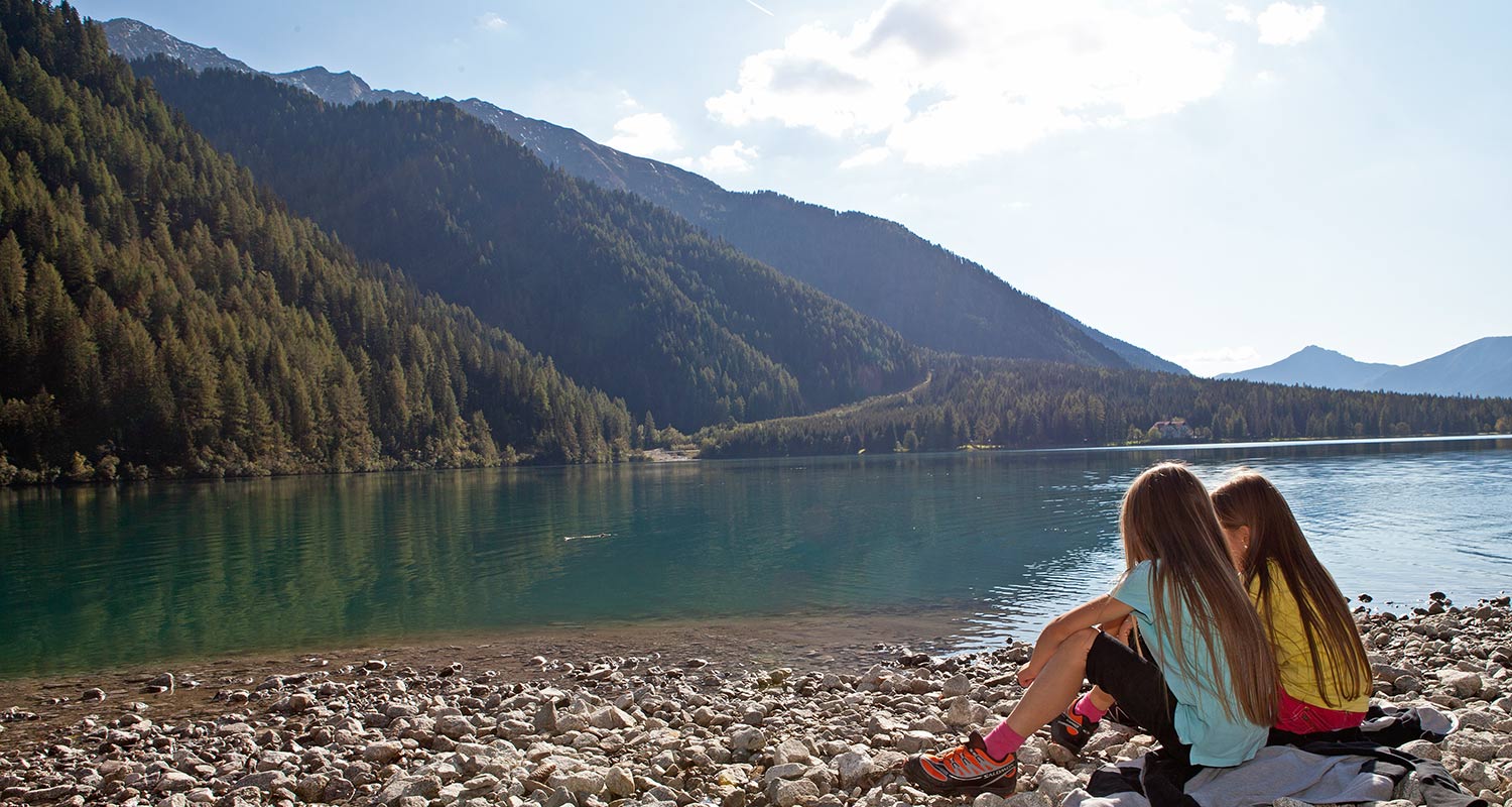 Zwei Mädchen sitzen am Ufer eines Sees mit Bergen im Hintergrund